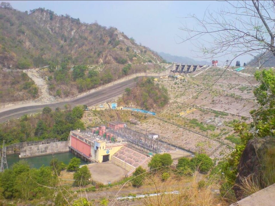 The Ramganga Dam complex at Kalagarh seen from a forested hillside: a red-and-cream powerhouse with an open switchyard beside a green tailrace pool, a long earth embankment sloping up behind it and a row of spillway gates set into the ridge in the distance