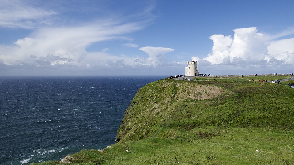 A bright green grassy headland at the Cliffs of Moher dropping in a sheer face to the dark blue Atlantic, with the round stone OBriens Tower and a line of tiny visitors along the clifftop path under piled white cumulus cloud