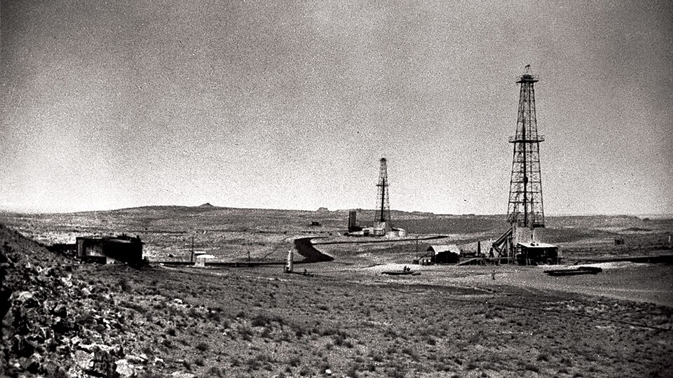A grainy black-and-white 1938 photograph of the Dammam oil field: two tall latticed steel drilling derricks standing on flat stony desert with low sheds, storage tanks and a dirt track between them, and a few tiny figures near the nearer rig