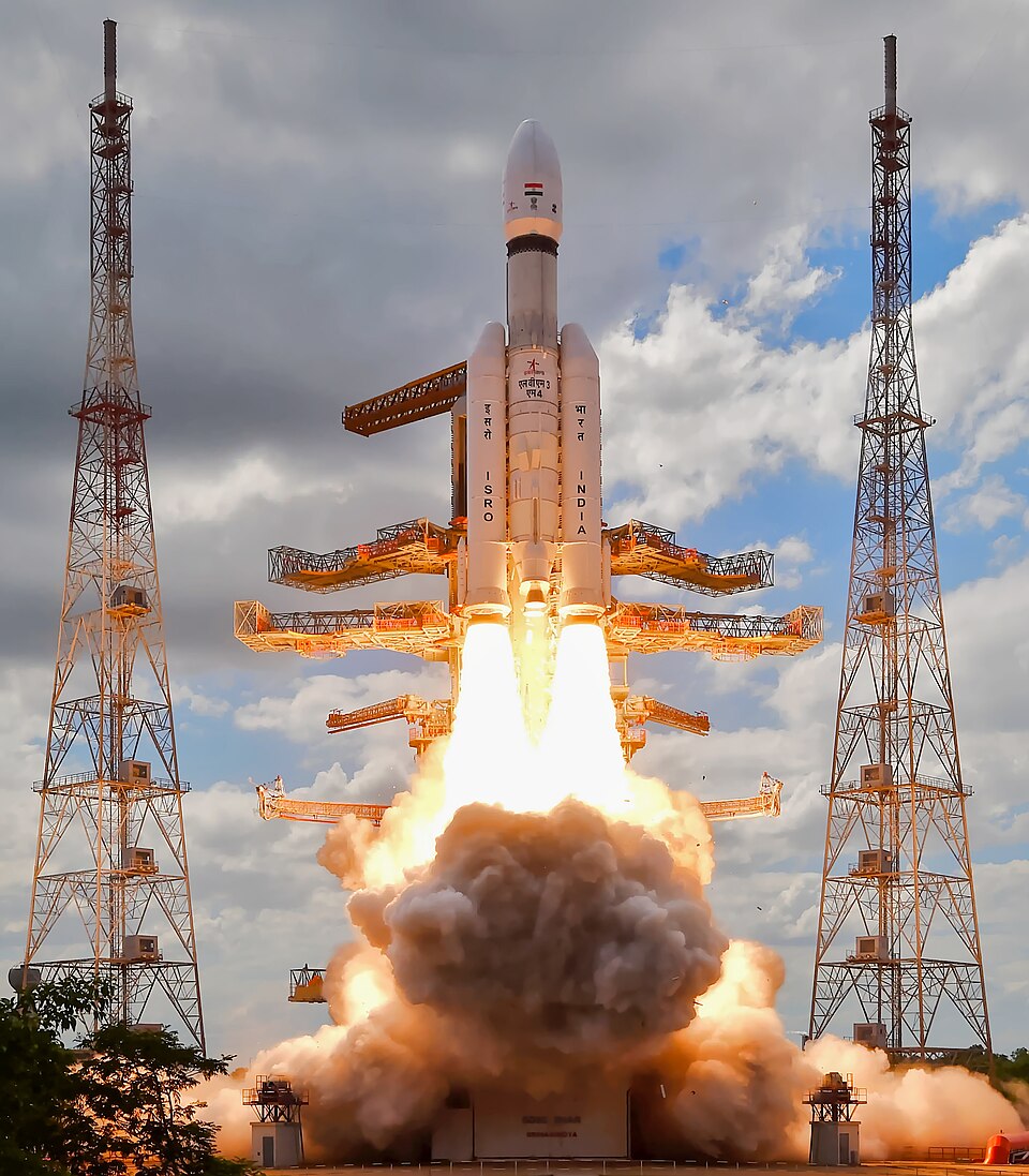 An LVM3 rocket lifting off between two lattice lightning towers at Sriharikota, its white payload fairing and nose cone at the top, the two strap-on boosters marked ISRO and INDIA throwing twin white flames, and a billowing orange-lit cloud of exhaust smoke spreading across the pad