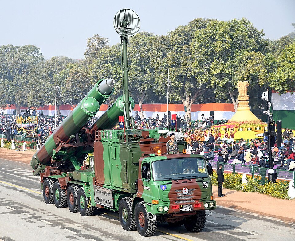 A camouflage-painted multi-axle military truck carrying a BrahMos mobile launcher along the Republic Day parade route in New Delhi, three long launch canisters raised at an angle behind the cab and a tall antenna mast beside them, with spectator stands and the national colours in the background