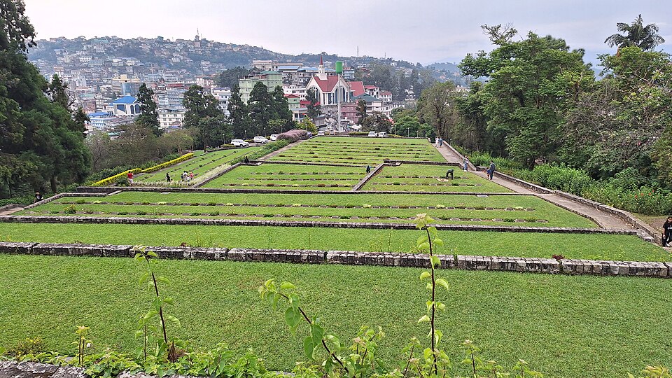 The terraced lawns of Kohima War Cemetery stepping down a hillside behind low stone retaining walls, each terrace set with long rows of small flat grave markers, with the houses of Kohima town and a church spire rising on the ridge beyond