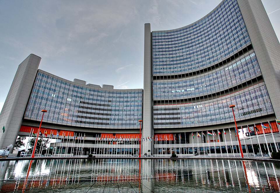 The curved glass-and-concrete office towers of the Vienna International Centre seen from across a still reflecting pool at dusk, with a long line of national flags on white poles standing in front of the orange-lit entrance level