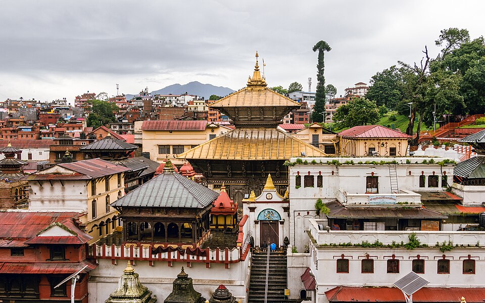 The two-tiered gilded pagoda roof and gold spire of the Pashupatinath temple rising above the whitewashed courtyards, carved wooden struts and grey-tiled shrine roofs of its complex, with the houses and green hills of the Kathmandu valley behind under a grey monsoon sky
