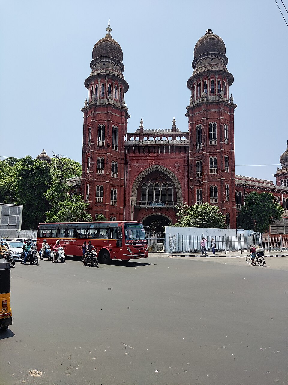 The deep red Indo-Saracenic facade of the Madras High Court in Chennai, with two tall towers capped by ribbed onion domes flanking a high pointed-arch entrance, seen from across a wide road with a red city bus, motorcycles and a cyclist passing in front