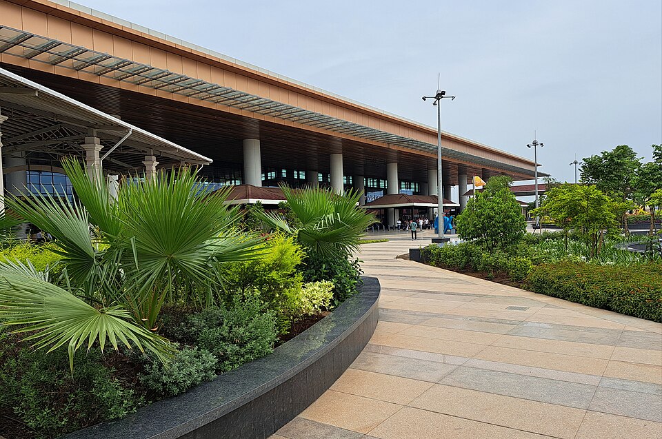 The kerbside frontage of a modern airport passenger terminal, a long slatted canopy on slender columns running away from the camera above the glass departures level, with fan palms in a curved planter and a wide granite forecourt in front