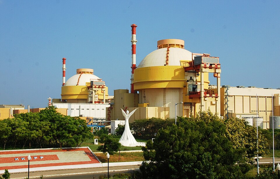The two domed reactor containment buildings of the Kudankulam nuclear power station under a clear sky, with red-and-white stacks alongside and landscaped grounds in front