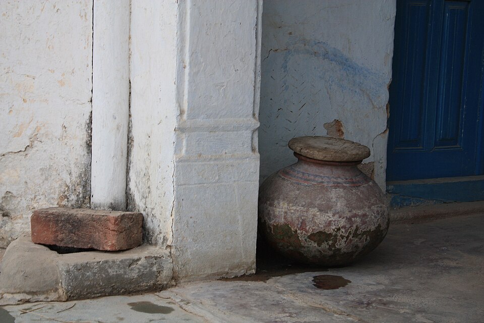 A large unglazed earthenware water matka with a flat clay lid standing on the floor beside a whitewashed doorway in an Indian village house, its lower half darkened by damp