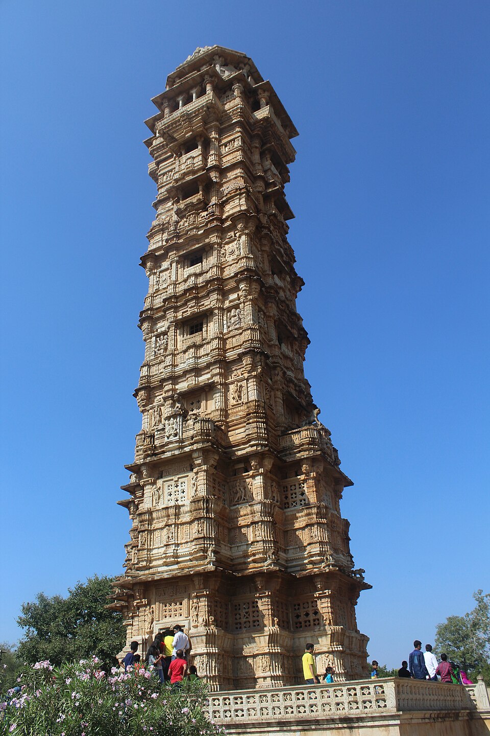 The nine-storeyed Vijay Stambha inside Chittorgarh Fort, a tall sandstone tower covered on every face with carved figures and pierced stone screens, seen from its base against a clear blue sky