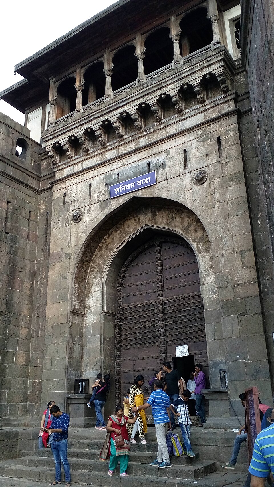 The Delhi Darwaza of Shaniwar Wada in Pune - a tall stone archway with heavy spike-studded wooden doors, a carved timber balcony above and a signboard reading 'शनिवार वाडा', with visitors on the steps