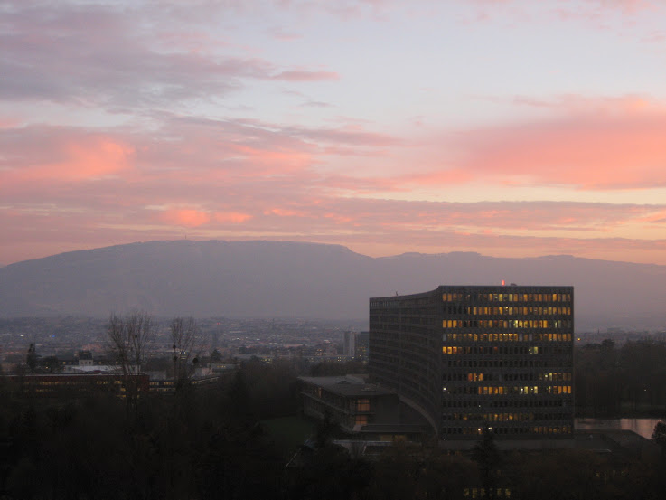 The long slab-shaped headquarters building of the International Labour Organization in Geneva, its windows lit at dusk, with the ridge of the Salève behind it under a pink sky