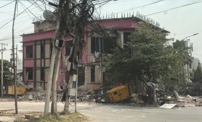 A pink two-storey building in Mandalay with its walls cracked open and its ground floor collapsed into rubble on the pavement, tangled overhead cables and a toppled roadside transformer in front of it