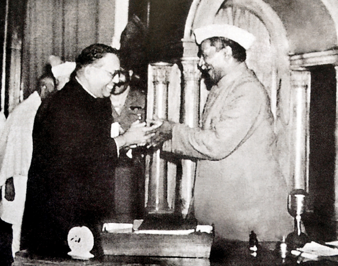 Black-and-white photograph of B. R. Ambedkar in a dark suit handing the bound final draft of the Constitution across a desk to Rajendra Prasad, who wears a white cap and sherwani, both smiling