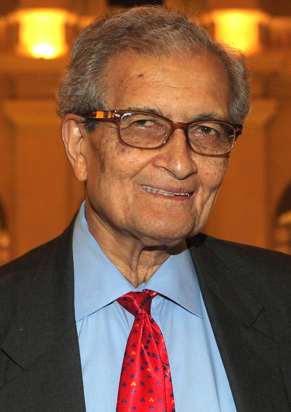 Head-and-shoulders portrait of Amartya Sen, grey-haired and wearing brown-rimmed glasses, a dark jacket, blue shirt and red patterned tie, smiling towards the camera