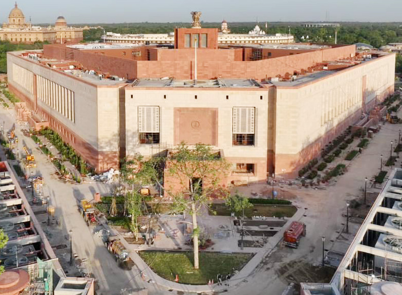 The new triangular Parliament House in New Delhi seen from above, its red-and-buff sandstone facades meeting at a corner and the national emblem sculpture standing on the roof