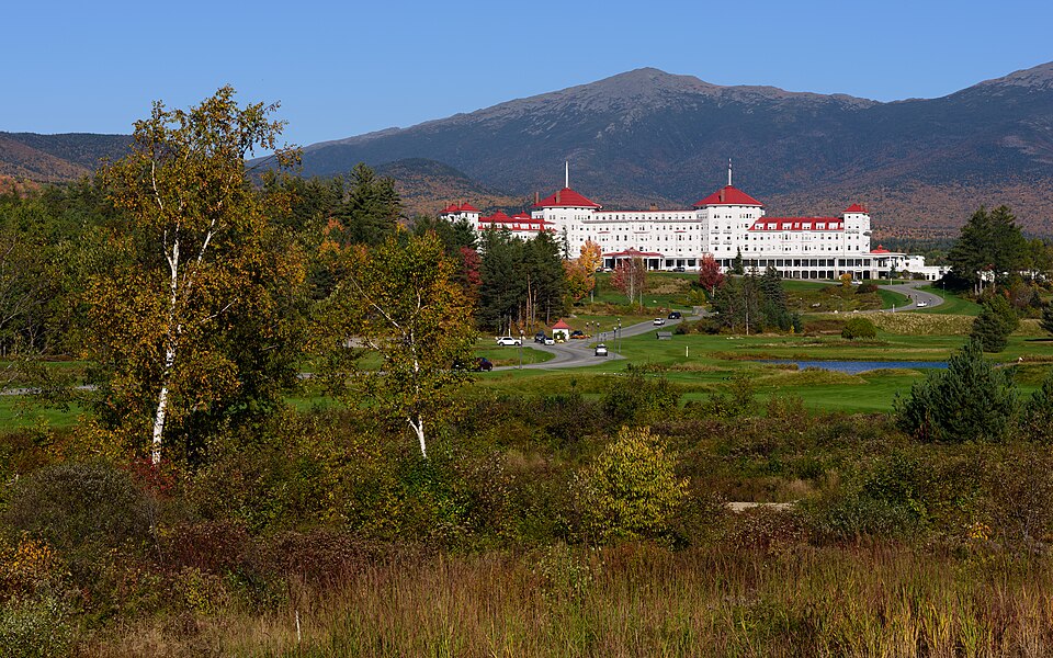 The white clapboard Mount Washington Hotel with its red roof and turrets standing on open lawns at Bretton Woods, with the wooded slopes of the White Mountains rising behind it
