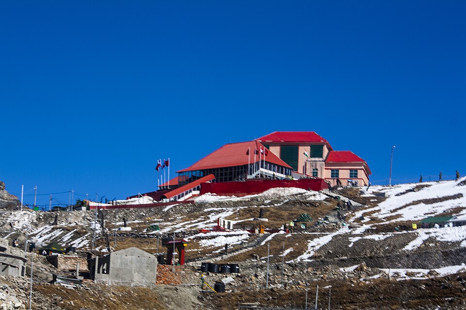 The red-roofed border post building on the crest of Nathu La, standing above snow-streaked rocky slopes crossed by fence lines and terraced tracks, under a deep blue sky