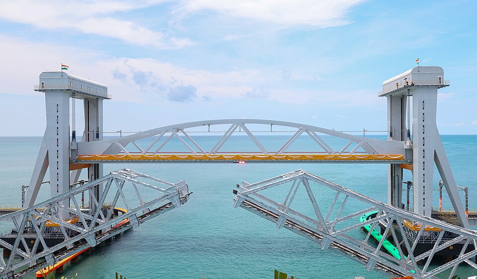 The New Pamban Bridge crossing the sea, its arched central span raised between two tall white lift towers, with the opened leaves of the old Pamban bascule bridge in the foreground