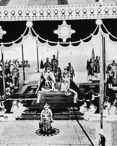Photograph of the 1911 Delhi Durbar: King George V and Queen Mary seated in robes and crowns on thrones on a carpeted dais beneath a decorated canopy, with attendants and guards ranged around them