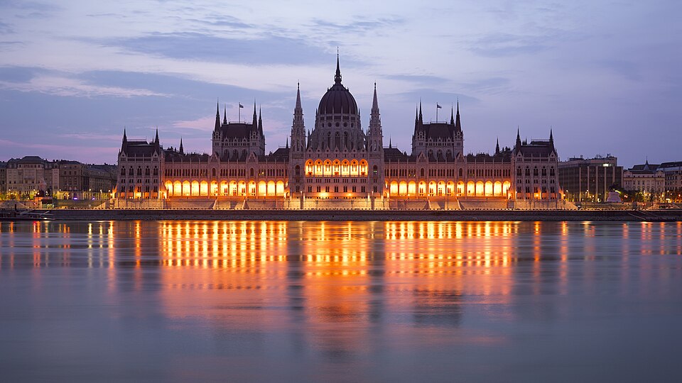 The Hungarian Parliament Building lit up at dusk on the Pest bank of the Danube, its long neo-Gothic facade, spires and central dome reflected in the still water