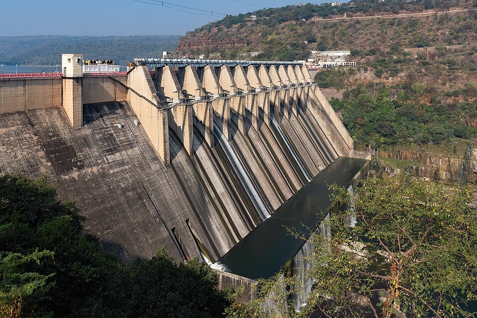 The Srisailam Dam seen from one abutment: a long concrete gravity dam with a row of crest gates and buttresses, water trickling down its face into the Krishna below, with wooded Nallamala hills on both banks