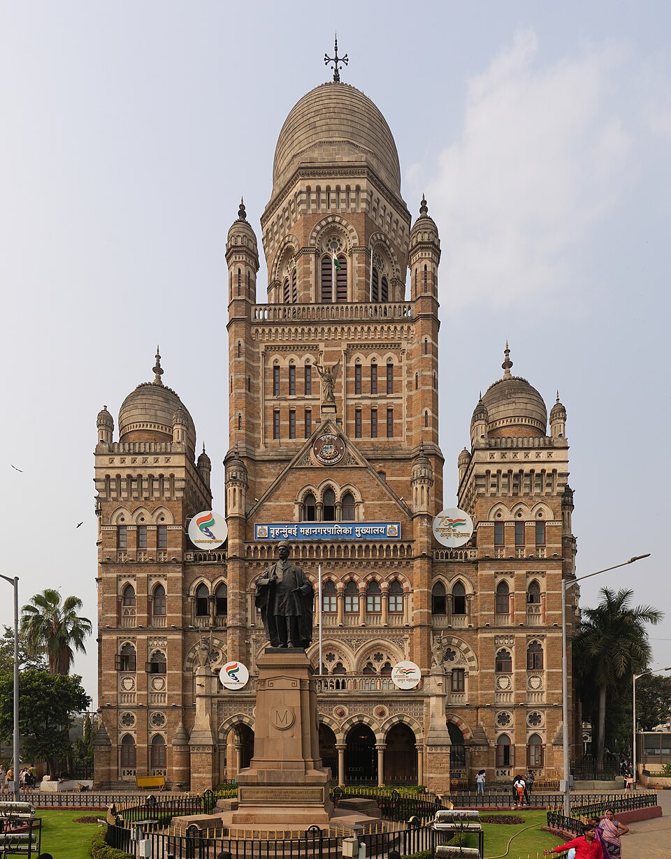 The Gothic Revival headquarters of the Brihanmumbai Municipal Corporation in daylight: a stone building with a central dome, flanking turrets and a gabled entrance, a Marathi nameboard across the facade and a statue on a plinth in front