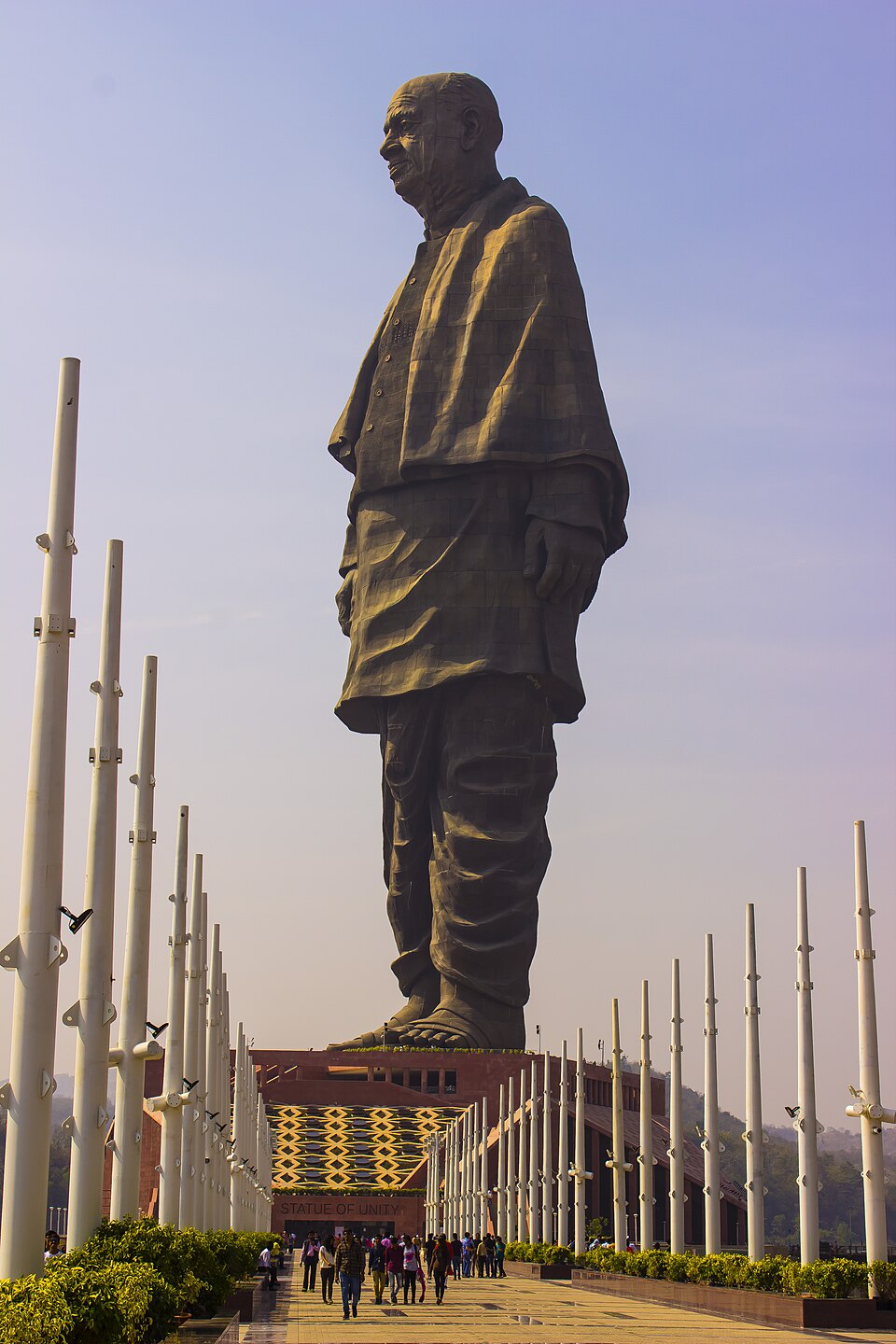 The Statue of Unity, a colossal bronze-clad standing figure of Sardar Vallabhbhai Patel on a red stone plinth, seen from the flagpole-lined approach walkway