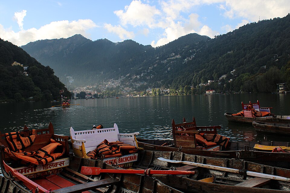 Wooden rowing boats with orange life jackets moored at the edge of Naini Lake, with the hillside town of Nainital and forested Kumaon ridges rising behind the water