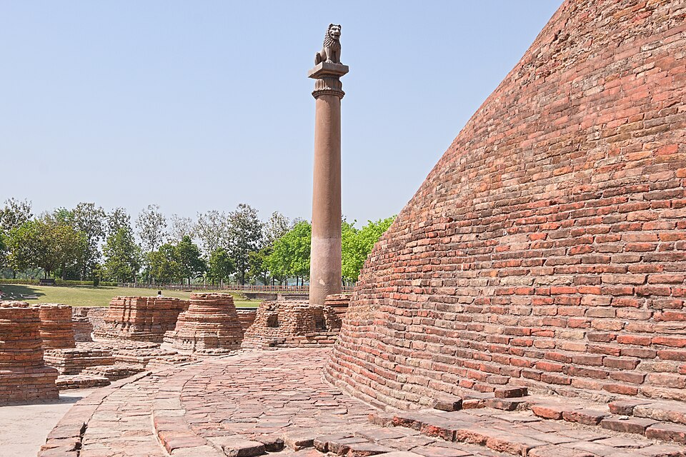 The polished monolithic sandstone Ashokan pillar at Kolhua, Vaishali, crowned by a single seated lion, standing beside the curved brick wall of the stupa