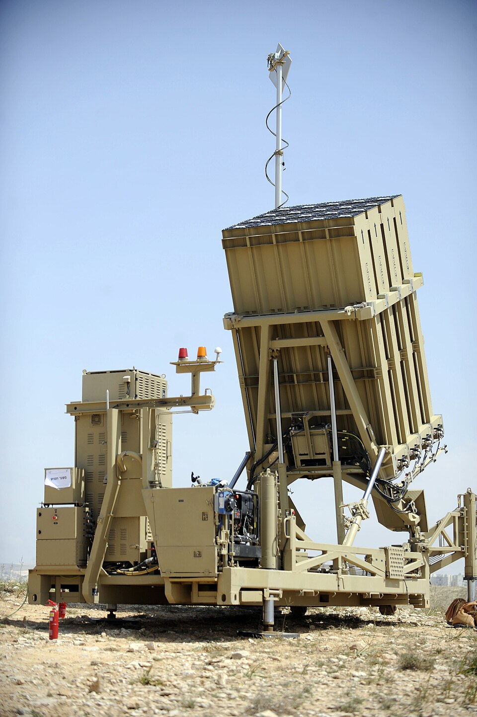 An Iron Dome launcher deployed on open stony ground - a boxy array of missile tubes tilted skyward on a sand-coloured towed trailer platform
