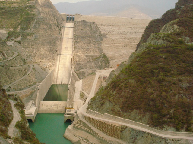 The Tehri Dam in its Himalayan gorge - a long concrete spillway chute running down the hillside beside the huge earth-and-rockfill embankment, with green water at its foot