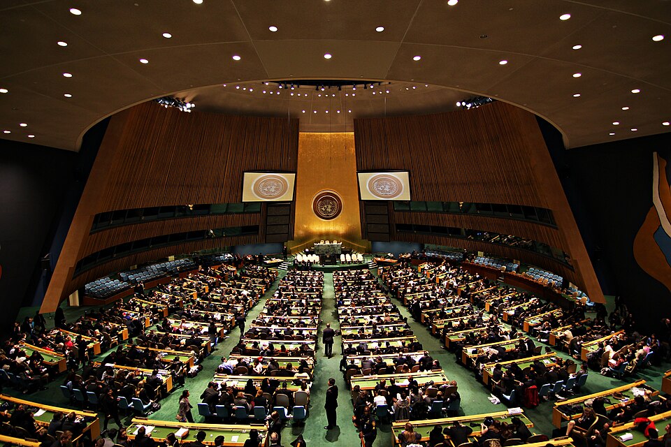 The United Nations General Assembly Hall in session, delegates filling curved rows of green desks below the gold rostrum wall bearing the United Nations emblem