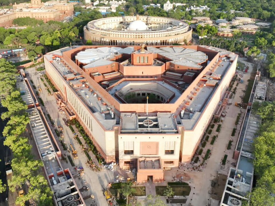 Overhead view of the new Parliament House in New Delhi, its triangular plan and sandstone courtyards clearly visible, with the circular old Parliament building behind it