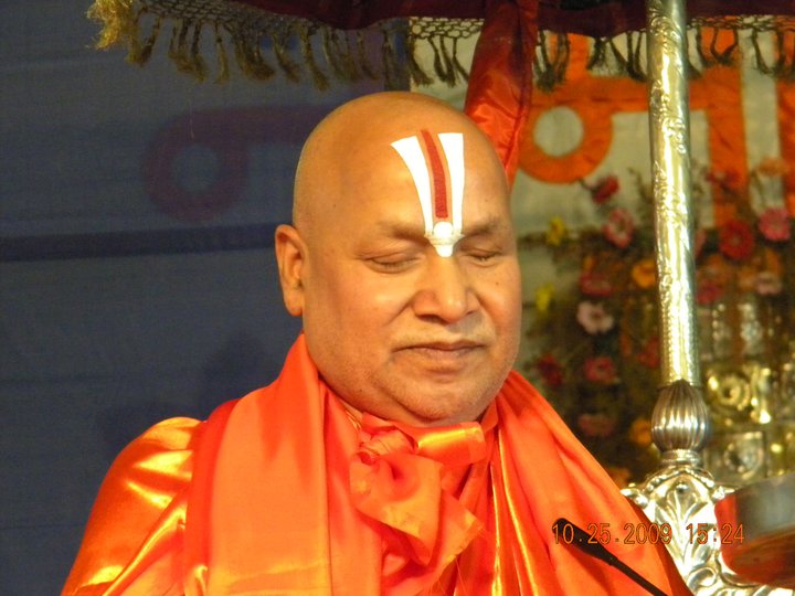Jagadguru Rambhadracharya, an elderly saffron-robed sage with a shaven head and a white-and-red Vaishnava tilak on his forehead, seated with his eyes closed beneath a decorated canopy