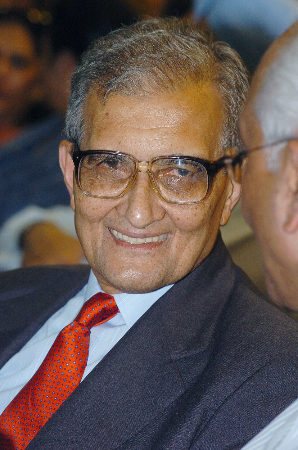 Amartya Sen, an elderly man with grey hair, thick-rimmed glasses, a dark suit and a red patterned tie, smiling towards the camera