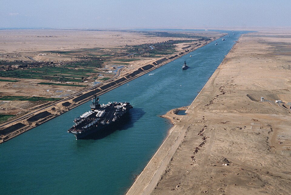 The Suez Canal running as a straight sea-level cut between desert banks, with a large aircraft carrier and other ships transiting in convoy