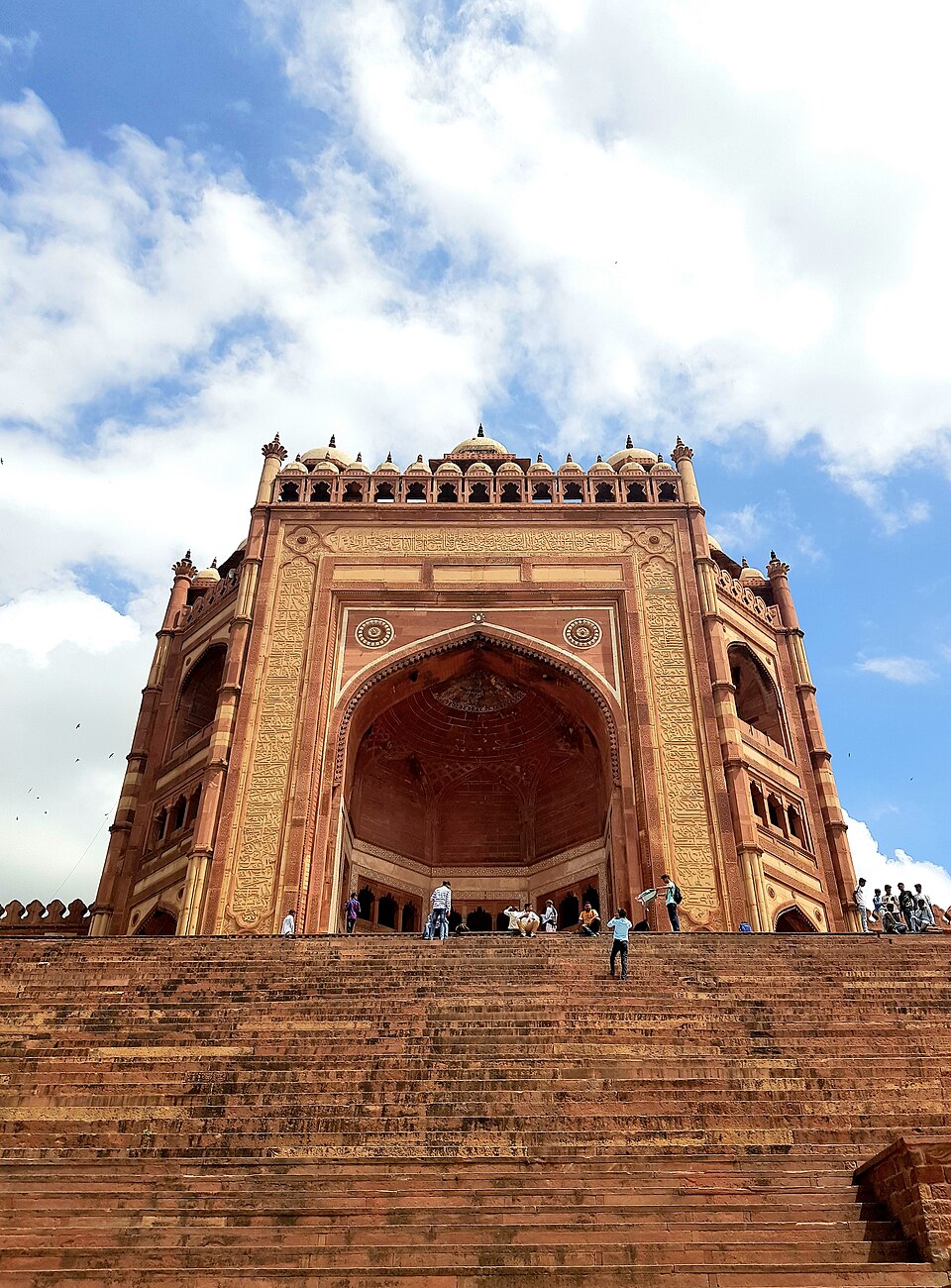 The Buland Darwaza at Fatehpur Sikri, a towering red-sandstone gateway with a deep arched recess, inscribed bands and a row of domed chhatris along the top, rising above a long flight of steps