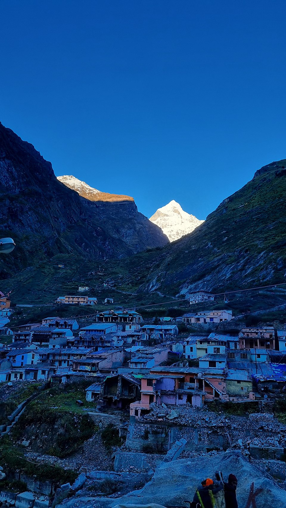 The stone and concrete houses of Mana village stacked on a steep Himalayan hillside at dusk, with a sunlit snow-covered peak rising between two dark ridges beyond