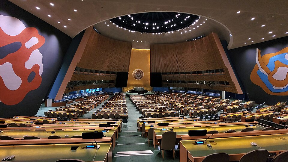 The United Nations General Assembly Hall seen from the back of the empty chamber: curved rows of desks facing the green marble rostrum and the gold UN emblem on the front wall