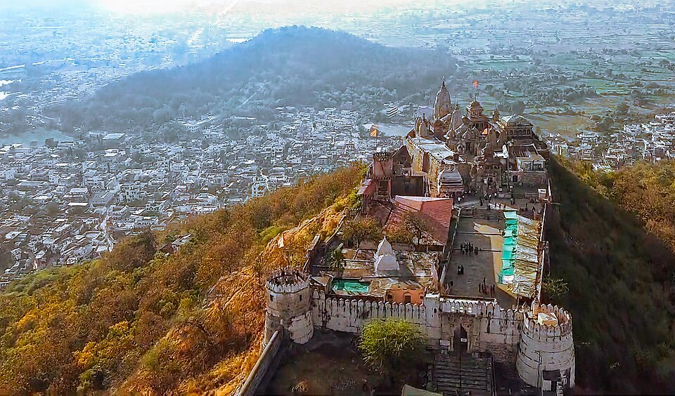 Aerial view of the fortified temple complex on Ramtek hill: crenellated stone ramparts and white shikharas along the crest of the ridge, with the town of Ramtek spread across the plain below